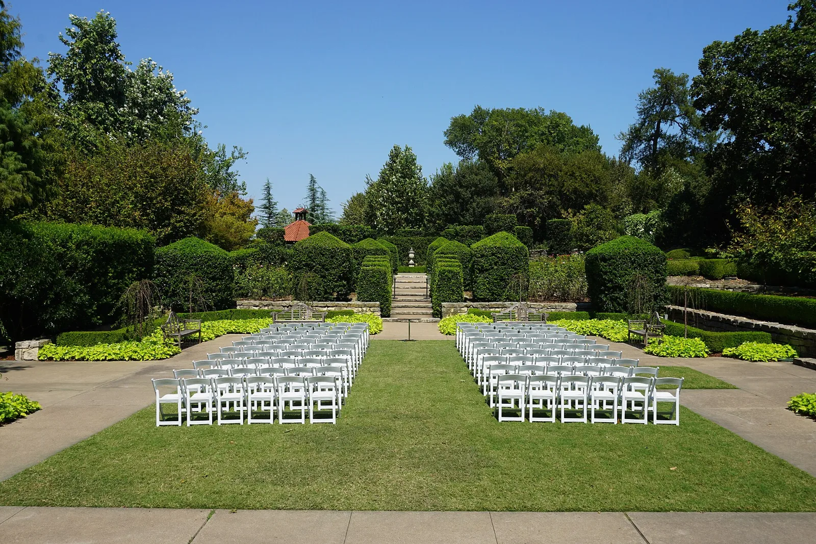 A sunken lawn ringed by clipped hedges and tall trees at the Dallas Arboretum, set with rows of white chairs
