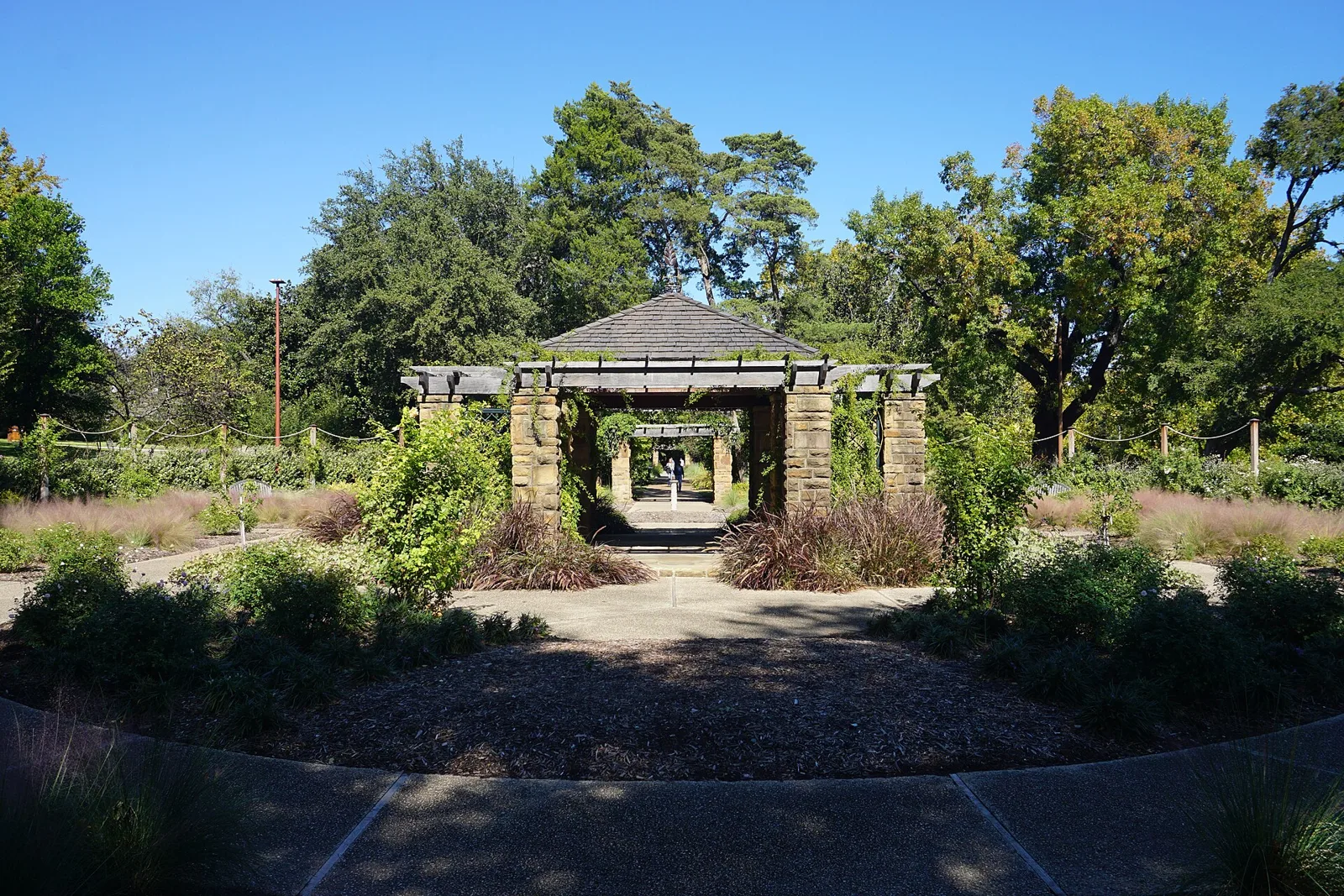 A stone and timber pergola framing a path through the Oval Rose Garden at the Fort Worth Botanic Garden