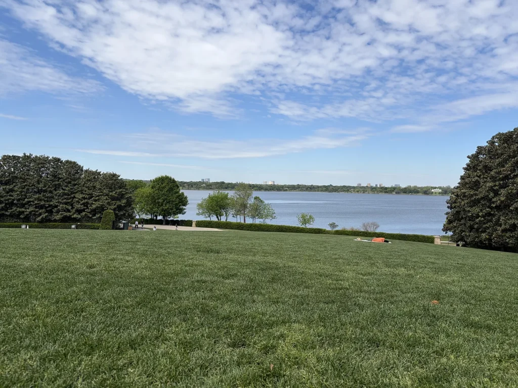 A wide green lawn at the Dallas Arboretum sloping down to White Rock Lake under a blue sky with clouds