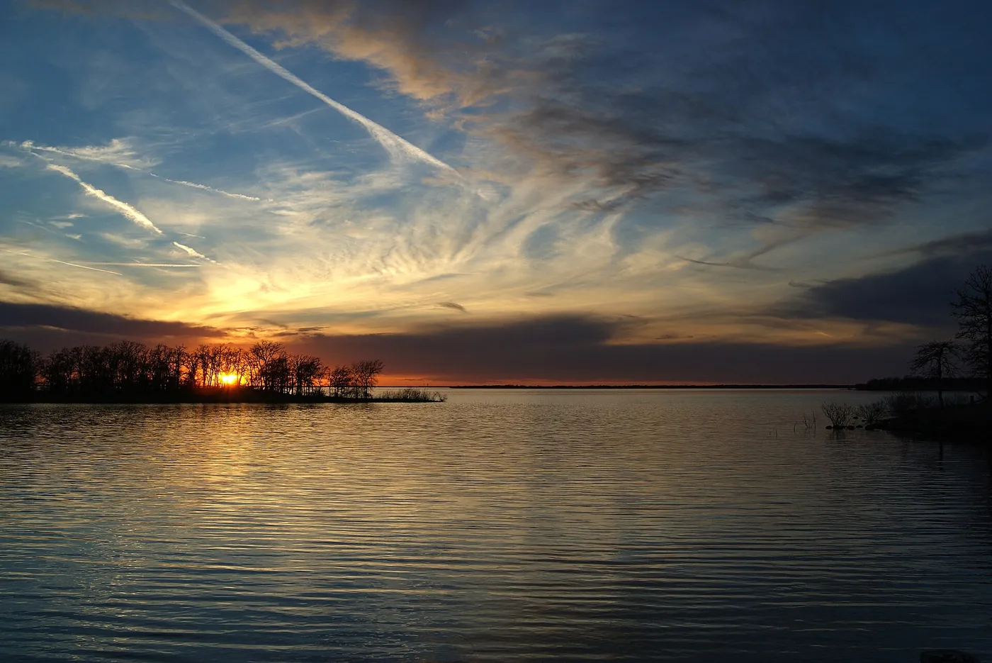 A low sun setting behind a line of trees across a calm lake, under streaked clouds